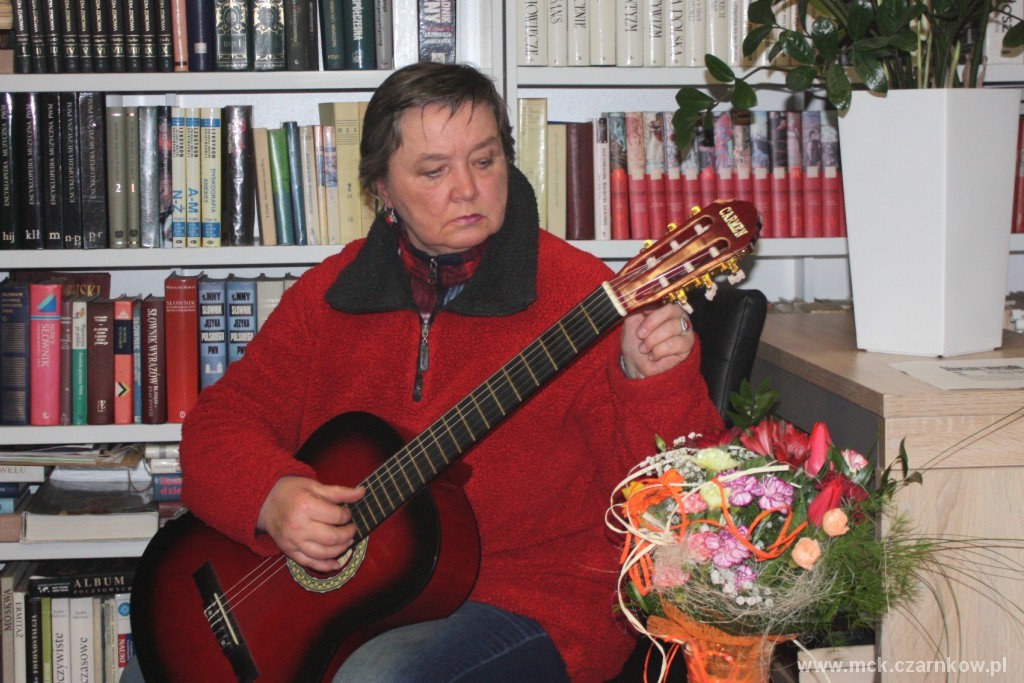 Kobieta strojąca gitarę, siedząca na&nbsp;tle regał&oacute;w z&nbsp;książkami