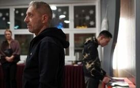 Na pierwszym planie stojący mężczyzna, na drugim rozmazana stojąca kobieta i mężczyzna przeglądający położone na stole pismo