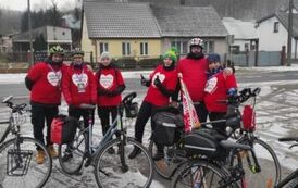 Grupa siedmiu os&oacute;b stojących z rowerami w koszulkch z nadrukiem serca wypełnionego napisem Wielka Orkiestra Świątecznej Pomocy na tle parterowych domk&oacute;w jednorodzinnych