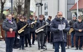 Mężczyzna m&oacute;wiący do mikrofonu na tle orkiestry dętej