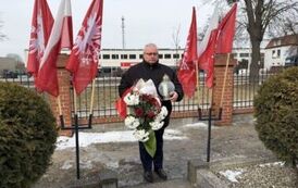 Mężczyzna stojący ze zniczem i wiązanką kwiat&oacute;w na tle biało-czerwonych flag i czerwonych flag z białym orłem