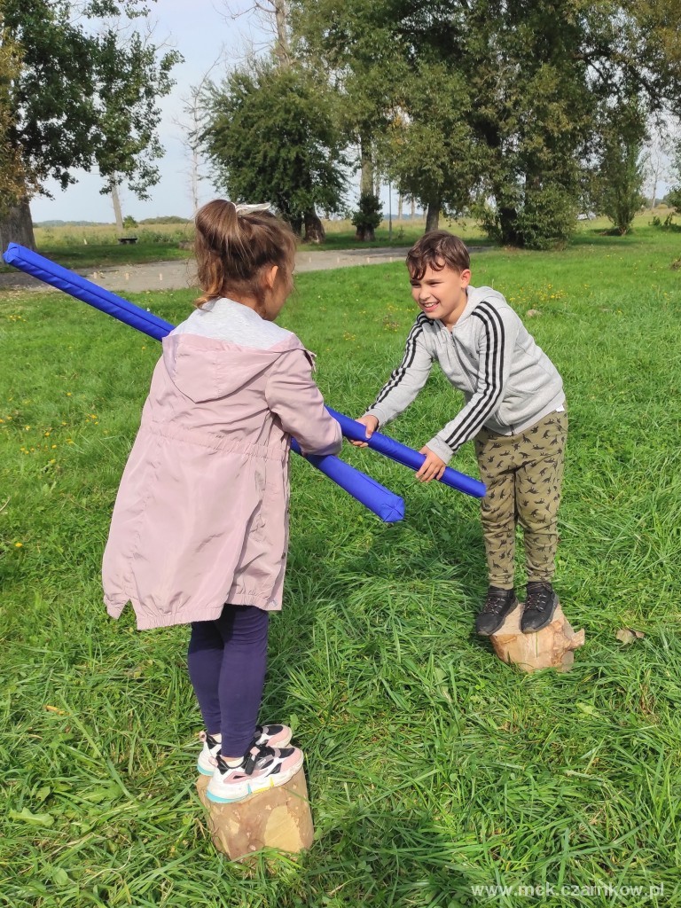 Dwoje dzieci stojąc na chybotliwych pieńkach walczy ze sobą piankowymi kijami