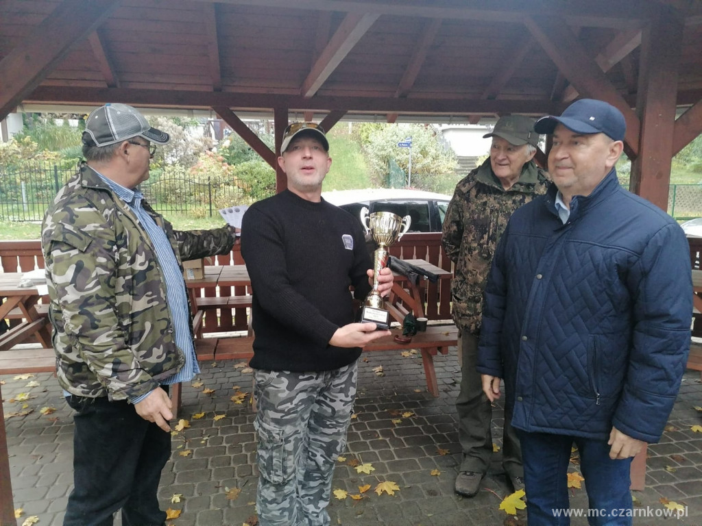 Czterech mężczyzn stojących pod dachem wiaty, jeden trzyma pozłacany puchar