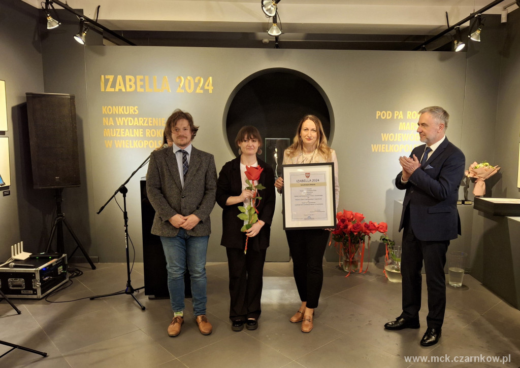 Dwie kobiety prezentujące kwiat i&nbsp;nagrodę w&nbsp;postaci zadrukowanej kartki w&nbsp;ramce, obok dw&oacute;ch mężczyzn