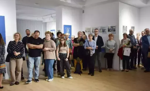 Ludzie róznej płci i wieku zgromadzeni w sali o iałych ścianach na których wisza ramy z fotografiami