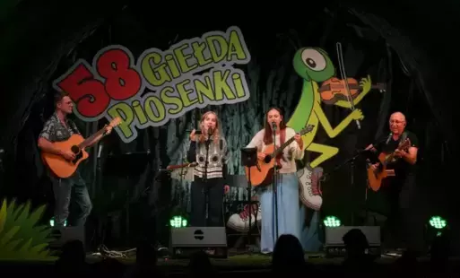 Dwóch mężczyzn z gitarami i dwie kobiety, jedna z gitarą, druga z marakasą, na scenie 