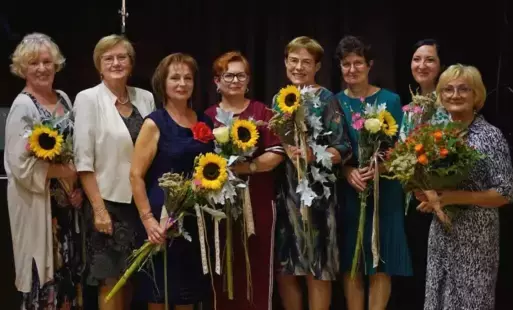 Na pierwszym planie brzeg pokryty rzadką roślinnością wodną, w tle parterowy domek z dwoma oknami i drzwiami