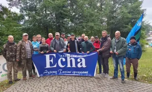 Grupa wędkarzy stojąca na tle drzew przed banerem z napisem Nadnoteckie Echa - Żyjemy Waszymi sprawami