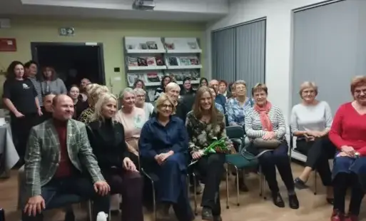 Lidzie siedzący na krzesłach, w tle ściana z regałem wypełnionym książkami