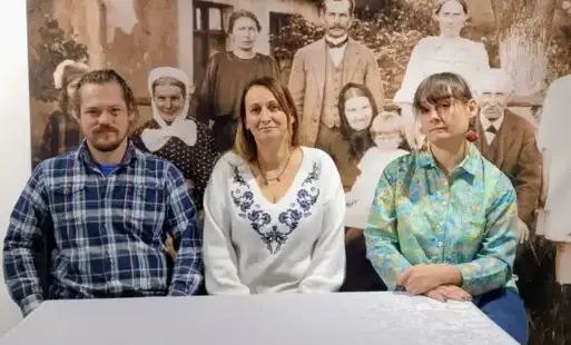 Dwie kobiety i mężczyzna siedzący na tle fototapety przedstawiającej fotografię w sepii z luźdzmi ustawionymi do wspólnego zdjęcia na tle chaty