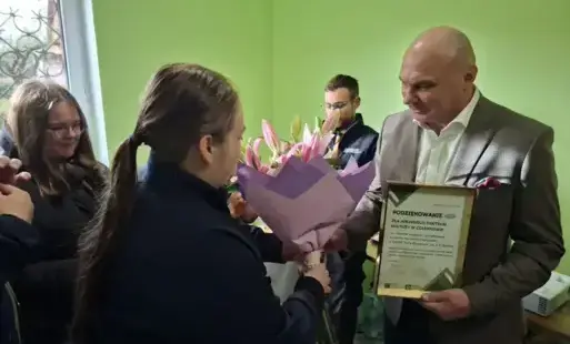 Mężczyzna trzymający w jednej ręce podziękowanie w ramce drugą ręką odbiera kwiaty
