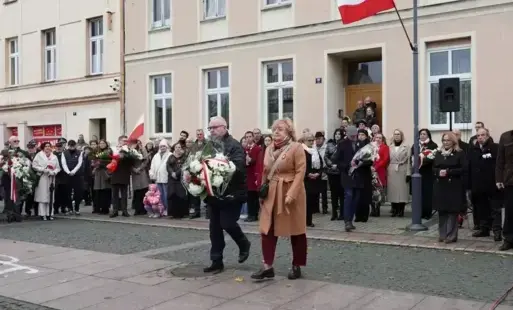 Idący mężczyzna z kwiatami, obok kobieta, w tle stojąca grupa ludzi, wielu trzyma kwiaty